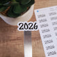 May include: A white sticker with the year 2026 written in black cursive font. The sticker is on a silver metal surface.