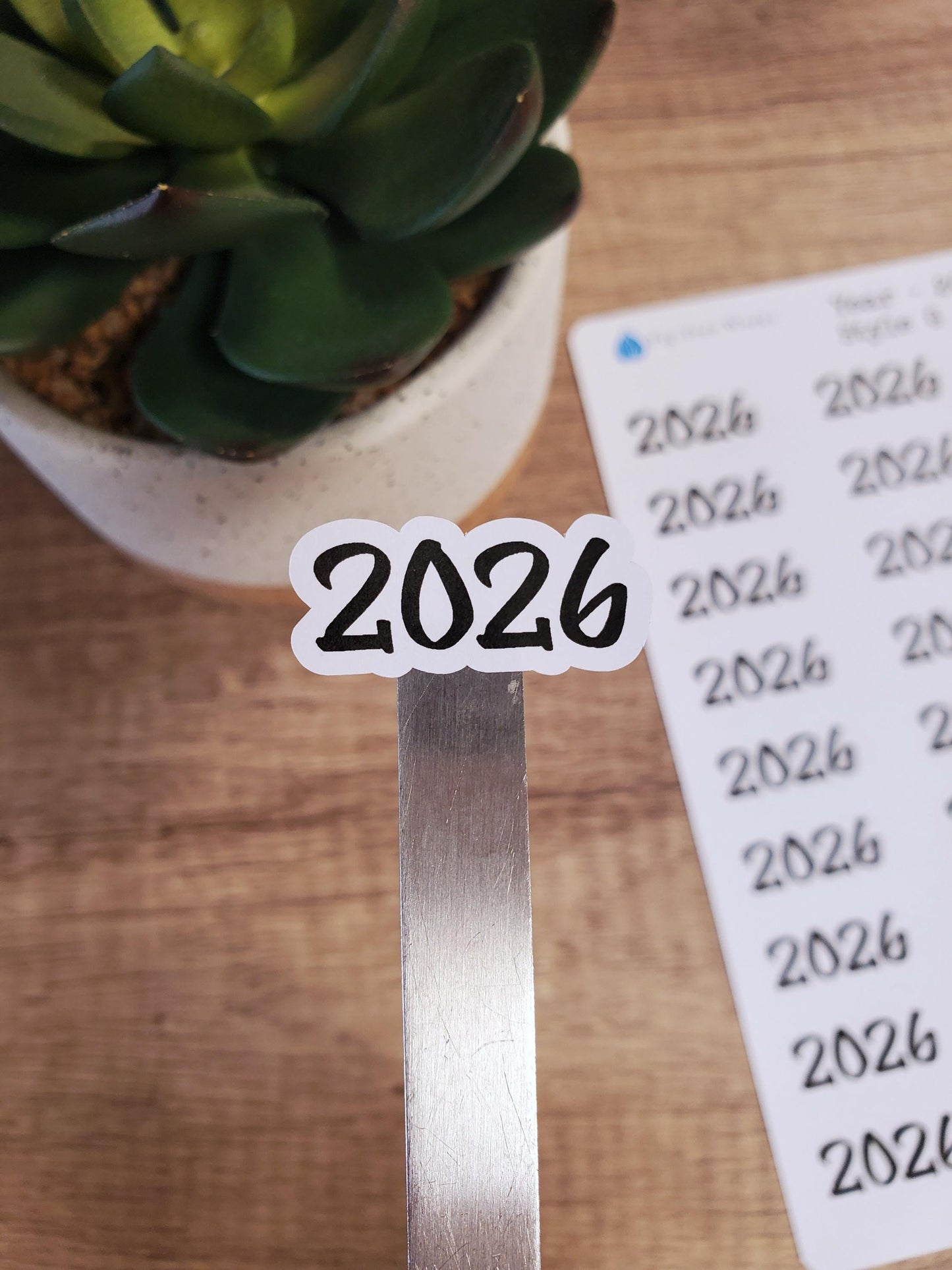 May include: A white sticker with the year 2026 written in black cursive font. The sticker is on a silver metal surface.