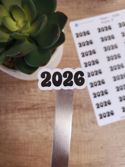 May include: A white sticker with the year 2026 in black, bubbly font. The sticker is on a silver metal surface.