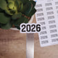 May include: A white sticker with the year 2026 printed in black. The sticker is placed on a silver metal rod. The sticker is next to a sheet of paper with the year 2026 printed multiple times.