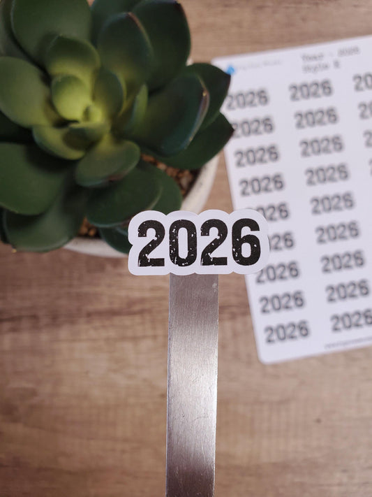 May include: A white sticker with the year 2026 printed in black. The sticker is placed on a silver metal rod. The sticker is next to a sheet of paper with the year 2026 printed multiple times.